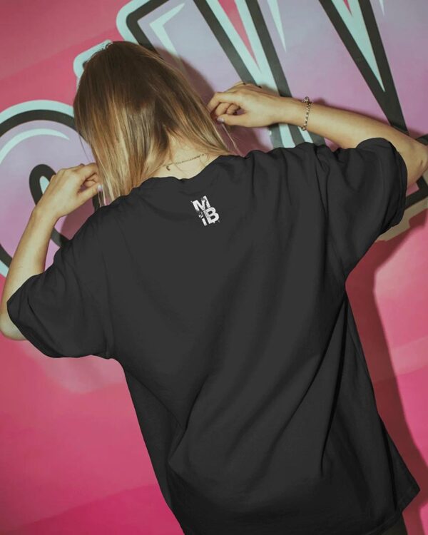 Person adjusting their hair while wearing a black T‑shirt with a small “MIB” back graphic, standing in front of a vibrant pink graffiti wall for a bold urban streetwear look