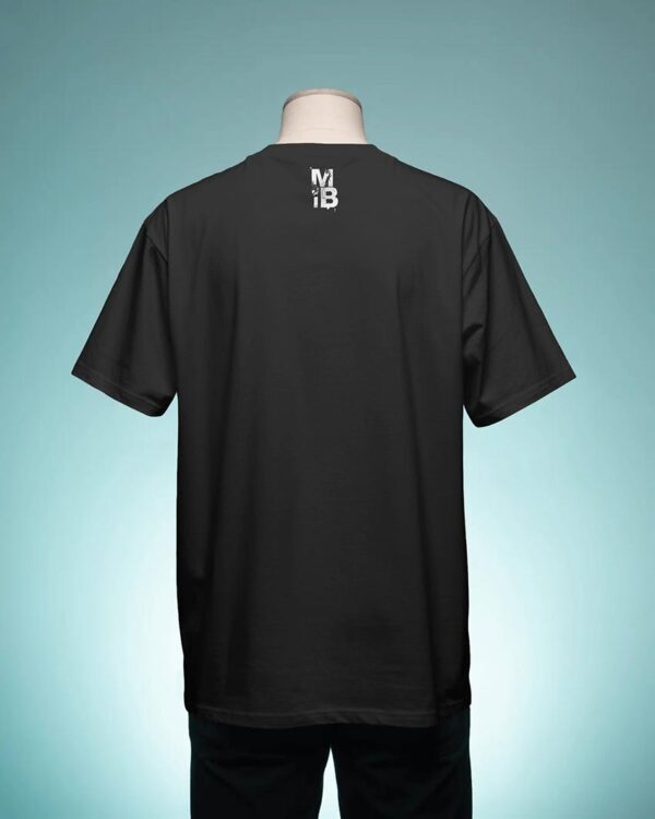 Person adjusting their hair while wearing a black T‑shirt with a small “MIB” back graphic, standing in front of a vibrant pink graffiti wall for a bold urban streetwear look