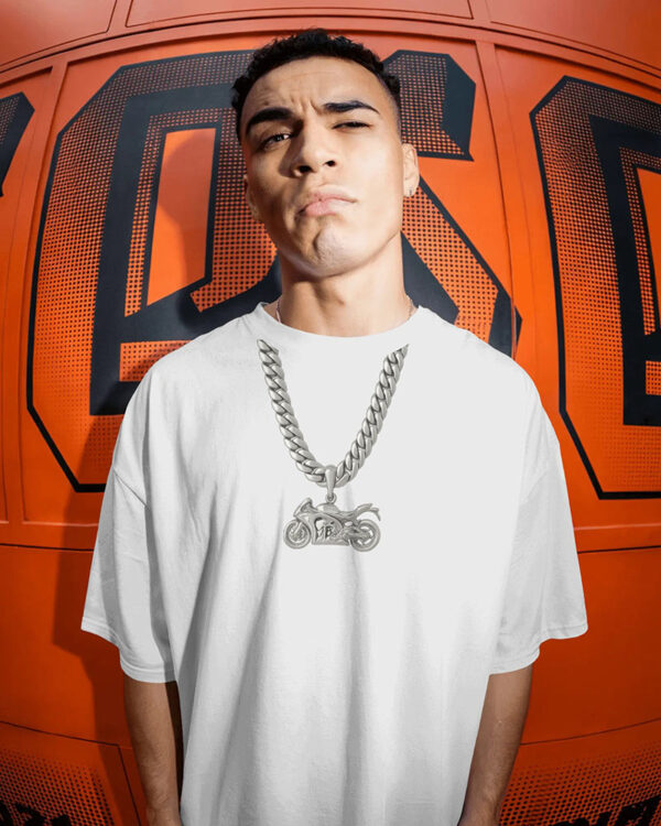 Person wearing an oversized white T‑shirt and a silver motorcycle pendant necklace standing in front of a bold orange wall with black graphic lettering, creating an urban streetwear aesthetic.