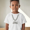 A young child standing indoors wearing a white T‑shirt with a printed silver chain and motorcycle pendant graphic, centered against a softly lit neutral background.