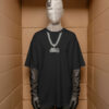 A mannequin wearing a black oversized T‑shirt with a printed silver chain and motorcycle pendant graphic, styled with black gloves and pants, standing inside a cardboard‑like display space.