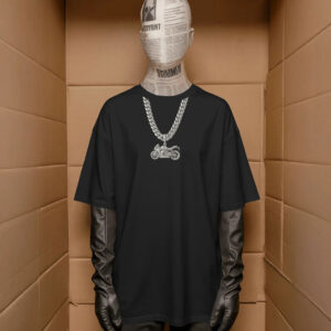 A mannequin wearing a black oversized T‑shirt with a printed silver chain and motorcycle pendant graphic, styled with black gloves and pants, standing inside a cardboard‑like display space.
