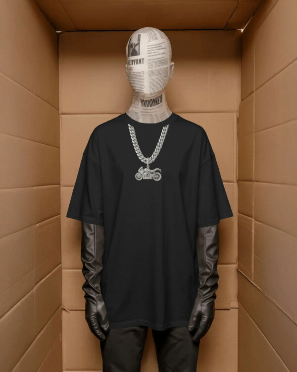 A mannequin wearing a black oversized T‑shirt with a printed silver chain and motorcycle pendant graphic, styled with black gloves and pants, standing inside a cardboard‑like display space.