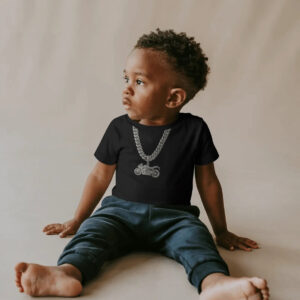 A young child wearing a black T‑shirt featuring a printed platinum chain and motorcycle pendant graphic, sitting barefoot on a light surface with a neutral background.