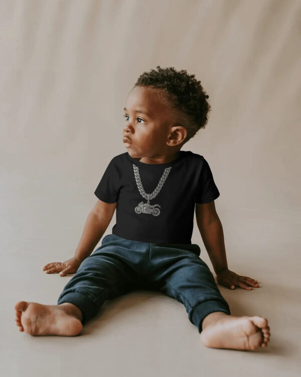 A young child wearing a black T‑shirt featuring a printed platinum chain and motorcycle pendant graphic, sitting barefoot on a light surface with a neutral background.