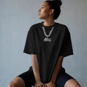 A person seated against a plain background wearing a black oversized T‑shirt featuring a printed silver chain and motorcycle pendant graphic, paired with black shorts.