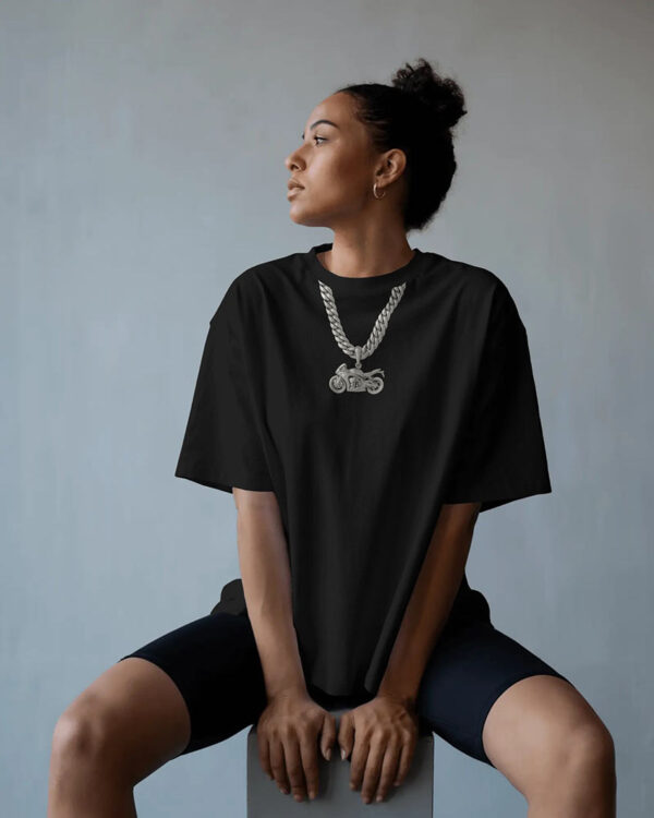 A person seated against a plain background wearing a black oversized T‑shirt featuring a printed silver chain and motorcycle pendant graphic, paired with black shorts.