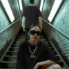 Two models in an urban stairwell wearing black streetwear graphic tees, one seated in oversized sunglasses and chain‑motorcycle print shirt, showcasing edgy moto‑inspired fashion in a dimly lit underground setting.