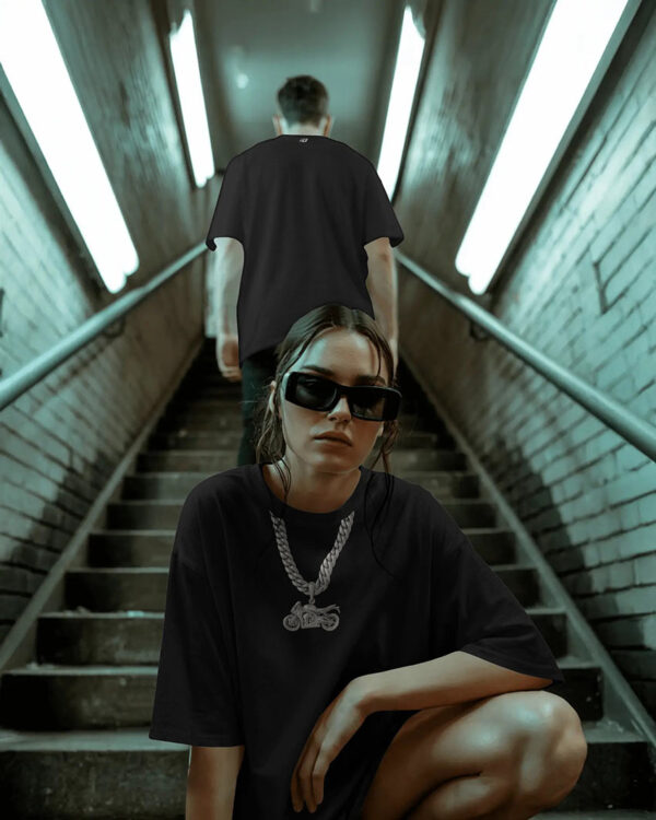 Two models in an urban stairwell wearing black streetwear graphic tees, one seated in oversized sunglasses and chain‑motorcycle print shirt, showcasing edgy moto‑inspired fashion in a dimly lit underground setting.