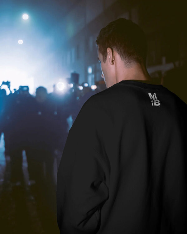 Person wearing a black MIB sweatshirt standing in a nighttime urban crowd, with phone lights and street haze illuminating the background