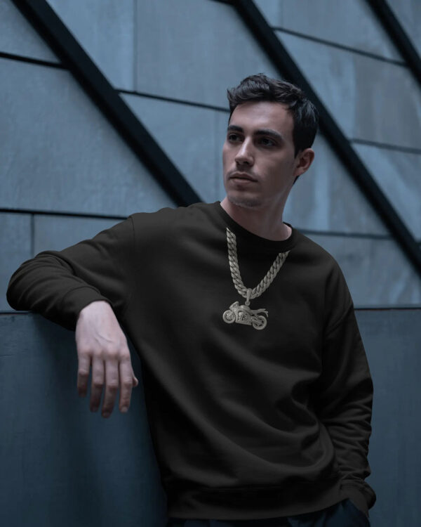 Man wearing a black sweatshirt with a gold motorcycle chain pendant, leaning against a blue wall in an urban industrial setting.