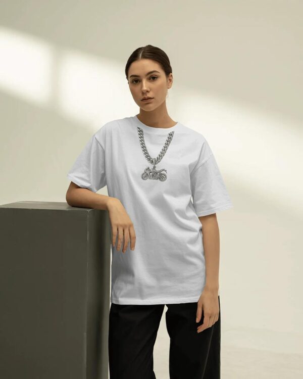 Person wearing a white T‑shirt with a bold printed chain-and-motorcycle pendant graphic, leaning against a block in a minimal studio setting for a clean urban streetwear look.
